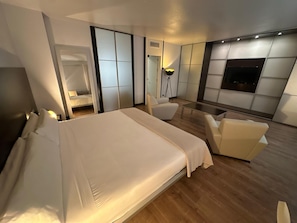 Una habitación de hotel moderna con una cama grande, un área de descanso con dos sillones, un escritorio y un ventanal amplio.