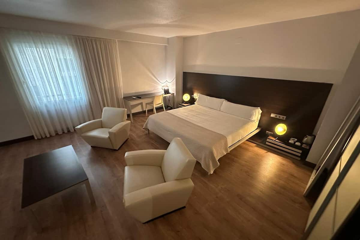 A hotel room with a bed, two armchairs, a desk, and a bedside table with lamps.
