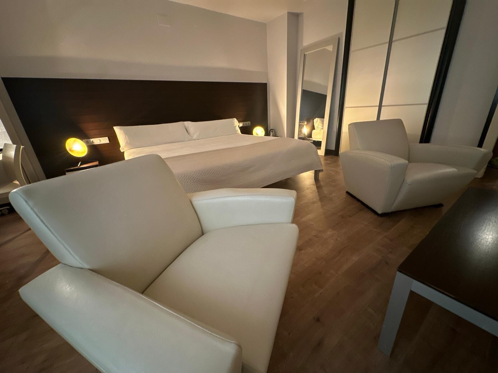 Una habitación de hotel moderna con una cama grande, dos sillones, una mesita y lámparas de noche.