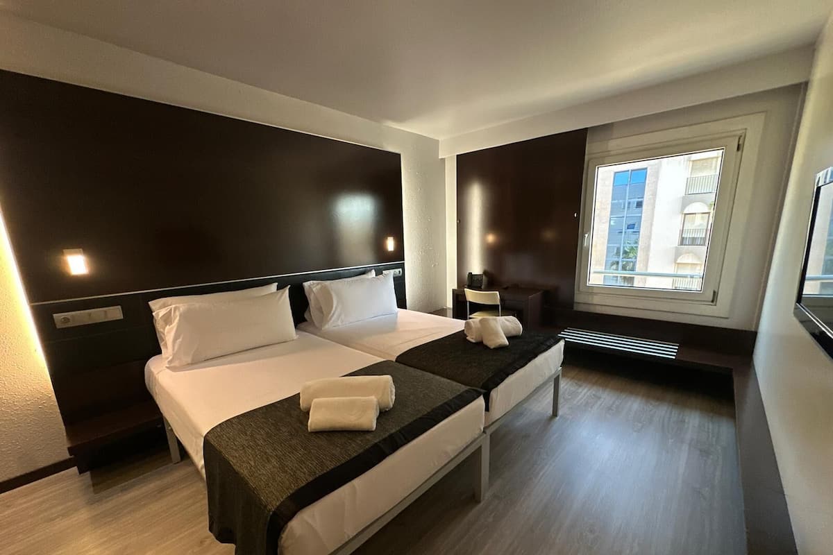 A hotel room with two beds, a desk, a chair, a window with a view of buildings, and a radiator.