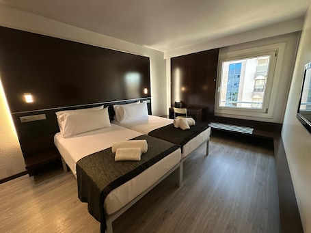A hotel room with two beds, a desk, a chair, a window with a view of buildings, and a radiator.. Puerta de Africa
