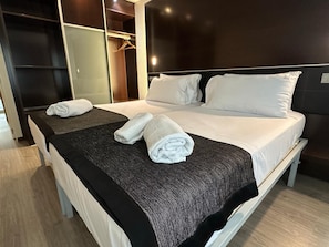 Una cama bien tendida con sábanas blancas, un plaid oscuro estampado y dos toallas enrolladas.