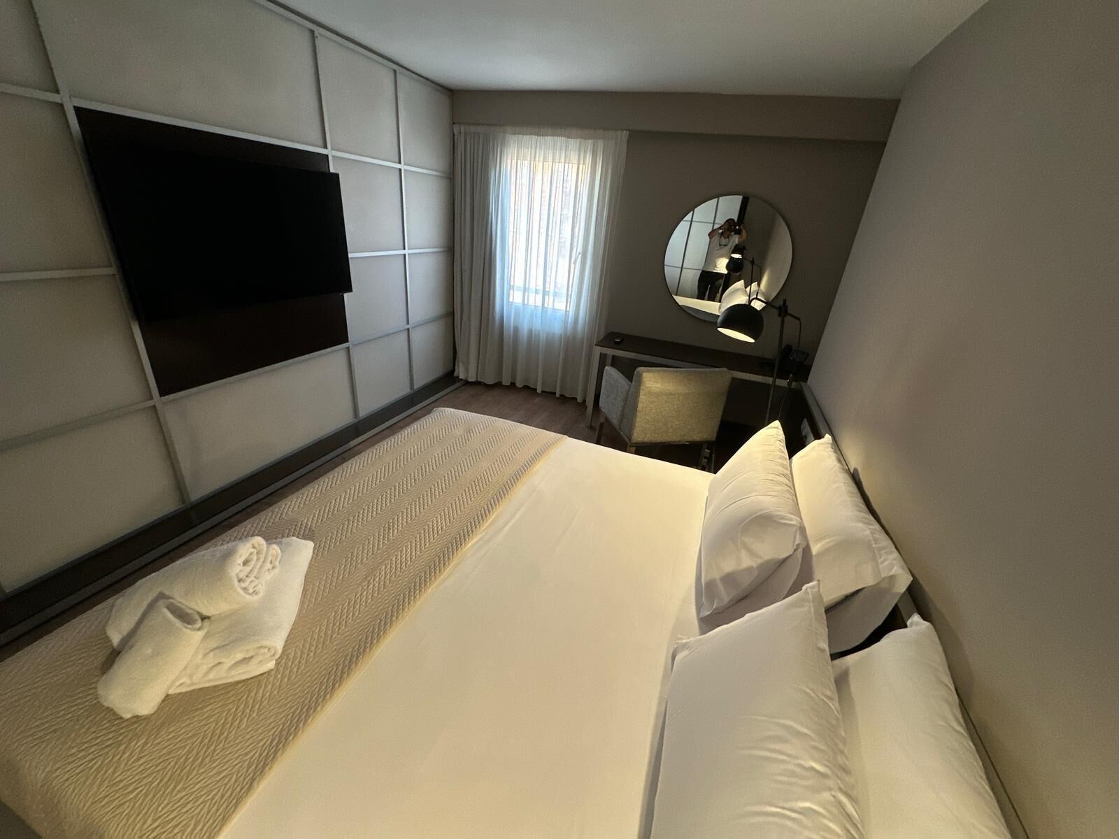 A hotel room with a bed, a desk, a chair, a TV mounted on the wall, and a large mirror.