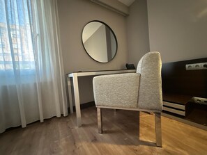 Une pièce moderne avec un plancher en bois, un fauteuil à revêtement gris, un bureau blanc et un miroir rond accroché au mur.