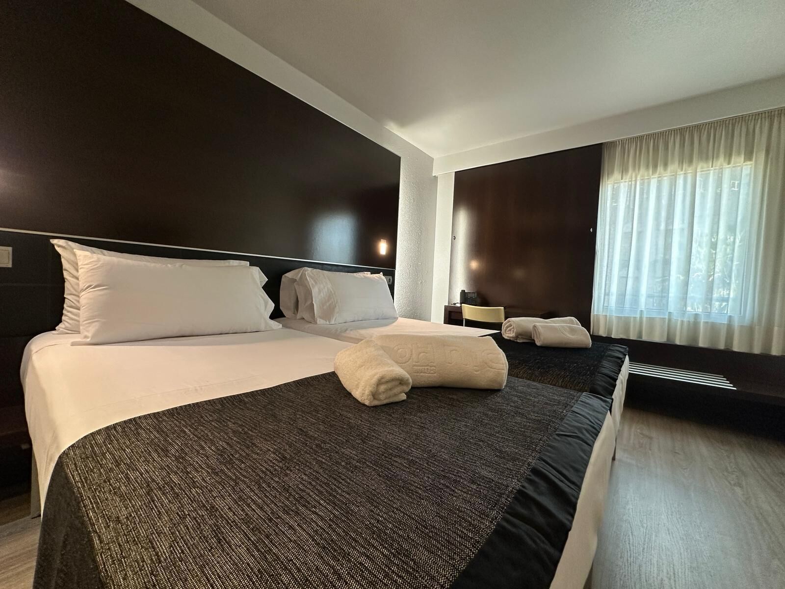 Ein Hotelzimmer mit einem großen Bett, einem Nachttisch, einem Sessel, einem Fenster mit Vorhängen und einer Wandlampe.