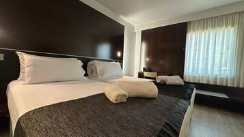 A hotel room with a large bed, a bedside table, a chair, a window with curtains, and a wall-mounted light.