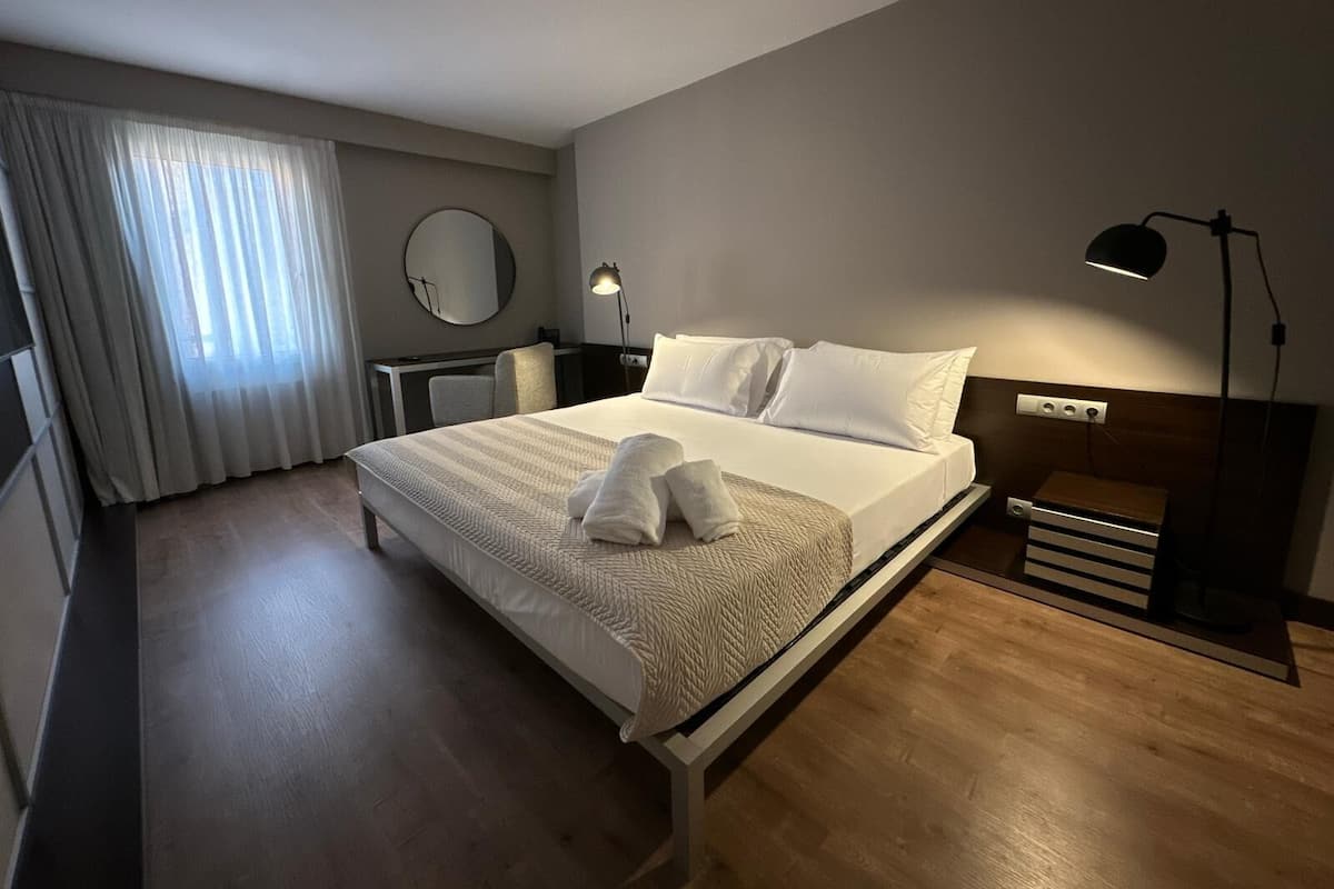 A hotel room with a bed, bedside lamp, desk, chair, and a mirror on the wall.