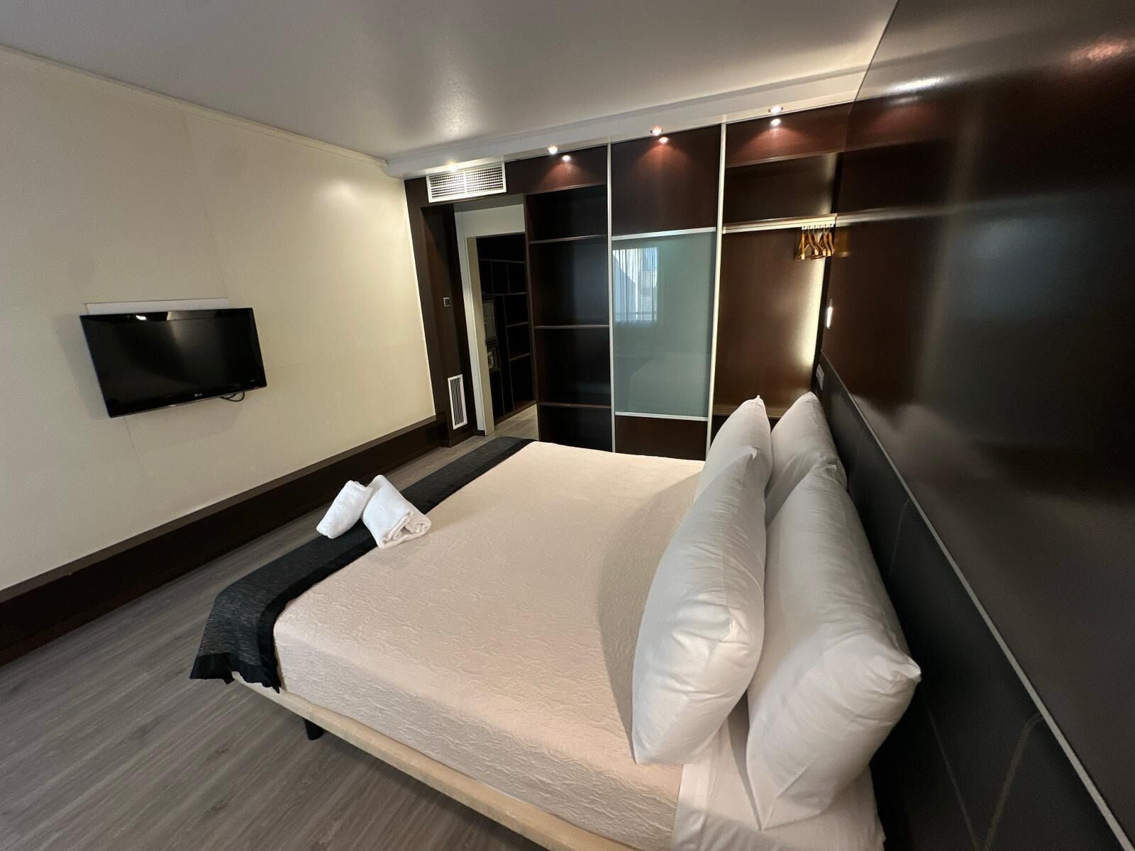 A modern hotel room with a large bed, a flat-screen TV, a walk-in closet, and a sliding glass door.