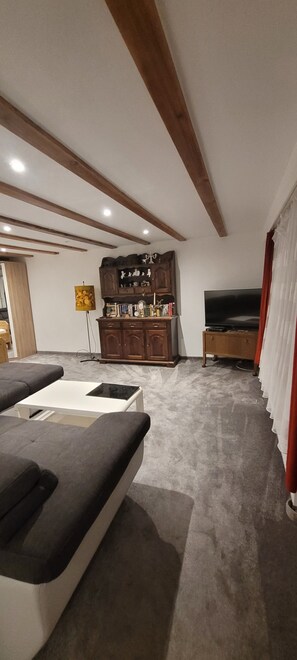 Ein Zimmer mit einer Couch, einem Fernseher, einer Kommode mit Dekoration und einer Decke mit Holzbalken.