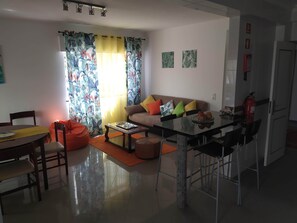 Ein Wohnzimmer mit einer Couch, einem Couchtisch und Sesseln, ein Essbereich mit Tisch und Stühlen sowie eine Küche mit Kühlschrank und Feuerlöscher.