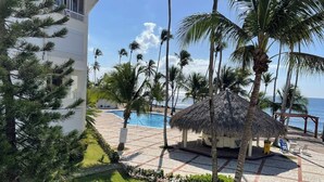 A pool area with a thatched gazebo, palm trees, and a view of the ocean. - Apartment in Albatros Club Resort, Juan Dolio (Playa Juan Dolio)