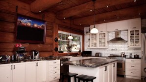 A kitchen with white cabinets, a large island, bar stools, a coffee machine, and a TV mounted on the wall. - Montagnard des Pays-d`en-Haut / Private Pool & Spa / Nature & Lake (Sainte-Adèle)