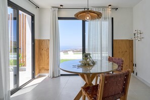 A modern dining area with a glass table, wooden chairs, a hanging light fixture, and a view through a glass door. - Mediterranean Window: Rural House with Sea Views, Wi-Fi & Air Conditioning (Torrox)