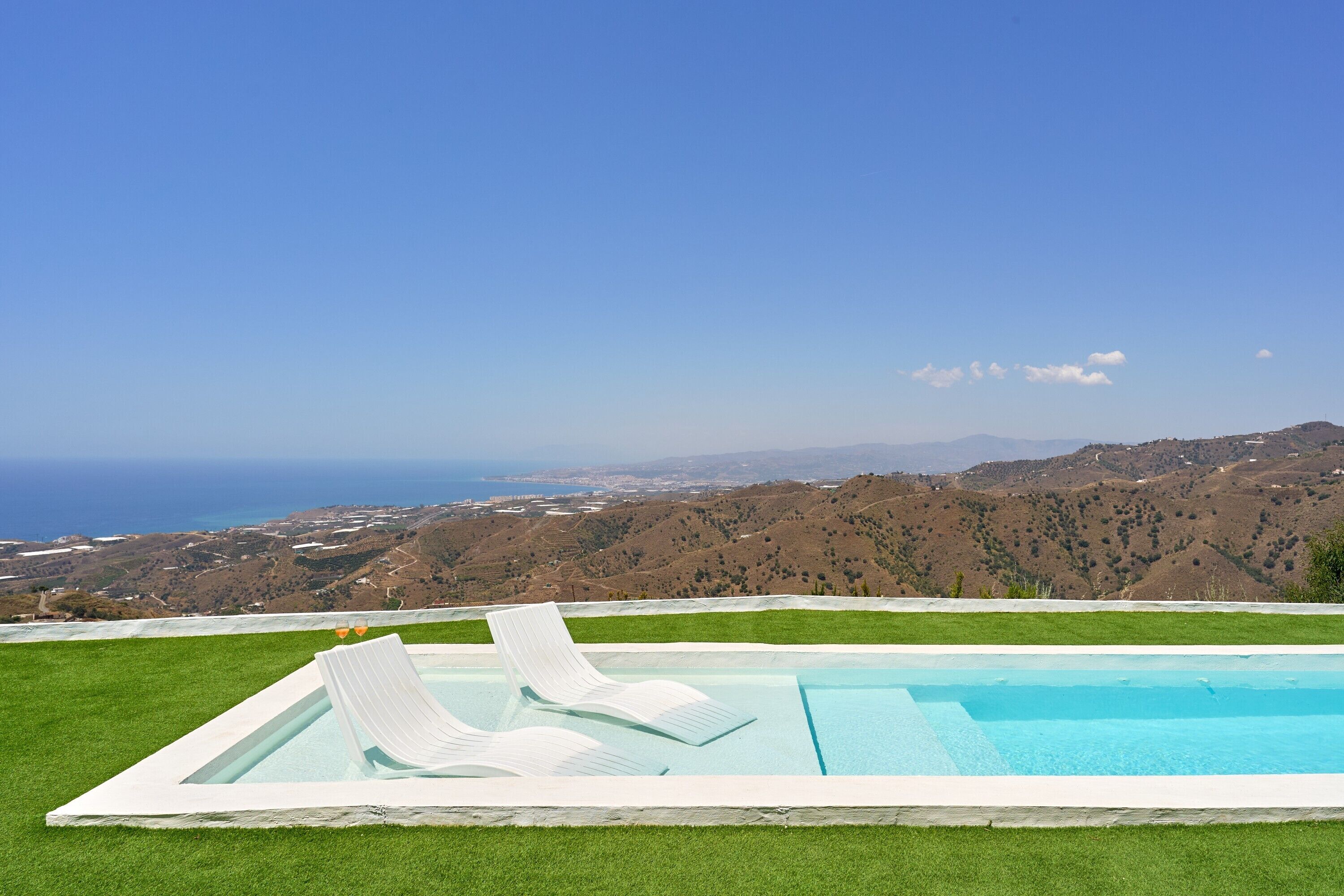 Uma piscina com duas espreguiçadeiras, cercada por grama verde, com vista para o oceano e colinas ao fundo.