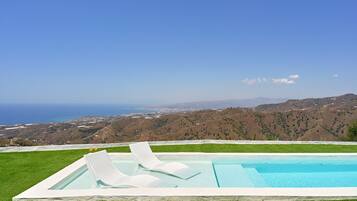 Uma piscina com duas espreguiçadeiras, cercada por grama verde, com vista para o oceano e colinas ao fundo.