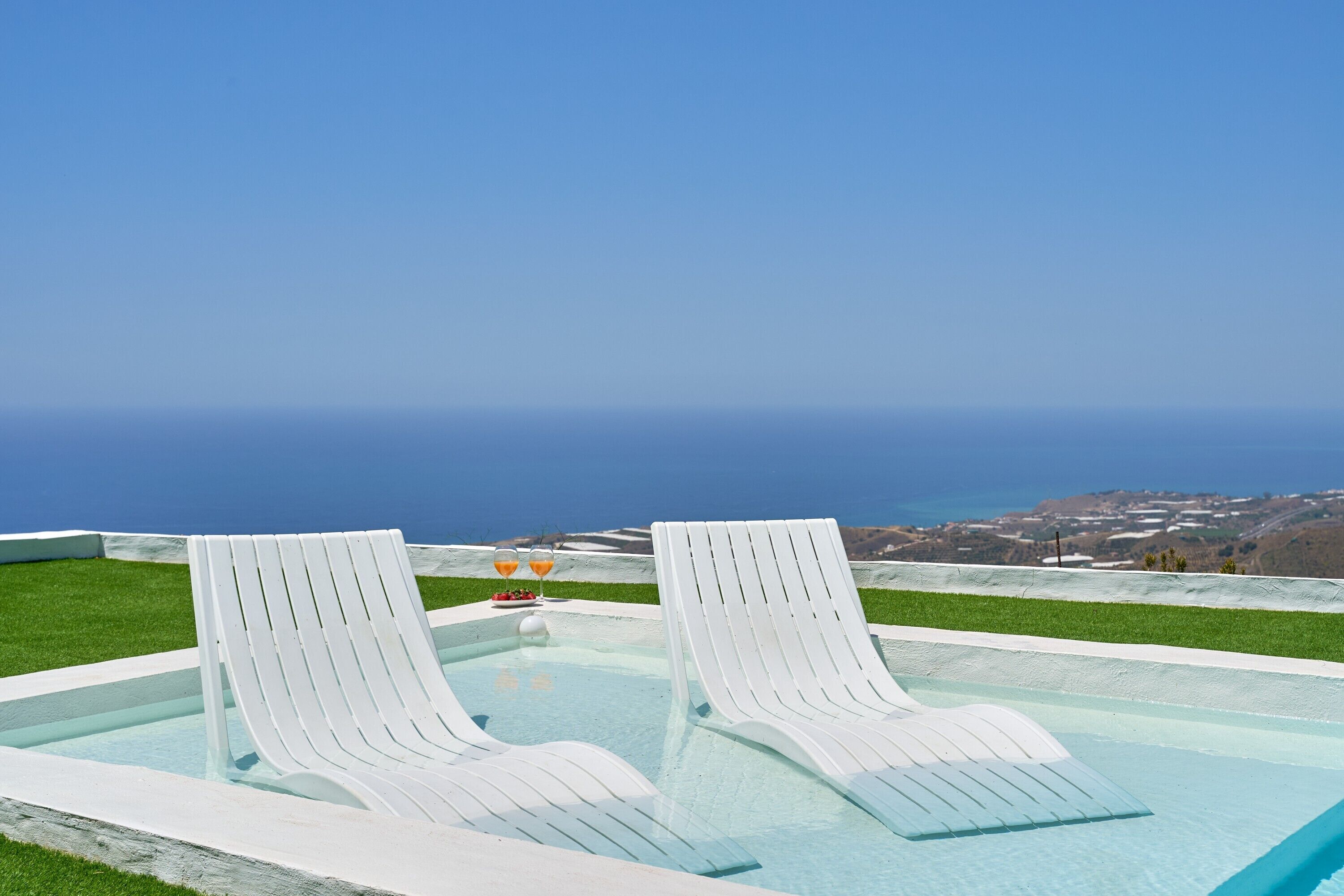 Une piscine bordée de deux chaises longues blanches et d’une table où sont disposées des boissons, le tout offrant une vue sur un paysage côtier.