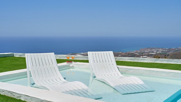 Una piscina con dos sillas de descanso blancas y una mesa con bebidas, con vista a un paisaje costero.