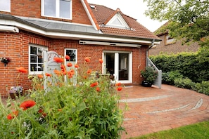 Une maison en briques avec un toit en tuiles, une porte d’entrée et un jardin où poussent des fleurs rouges.