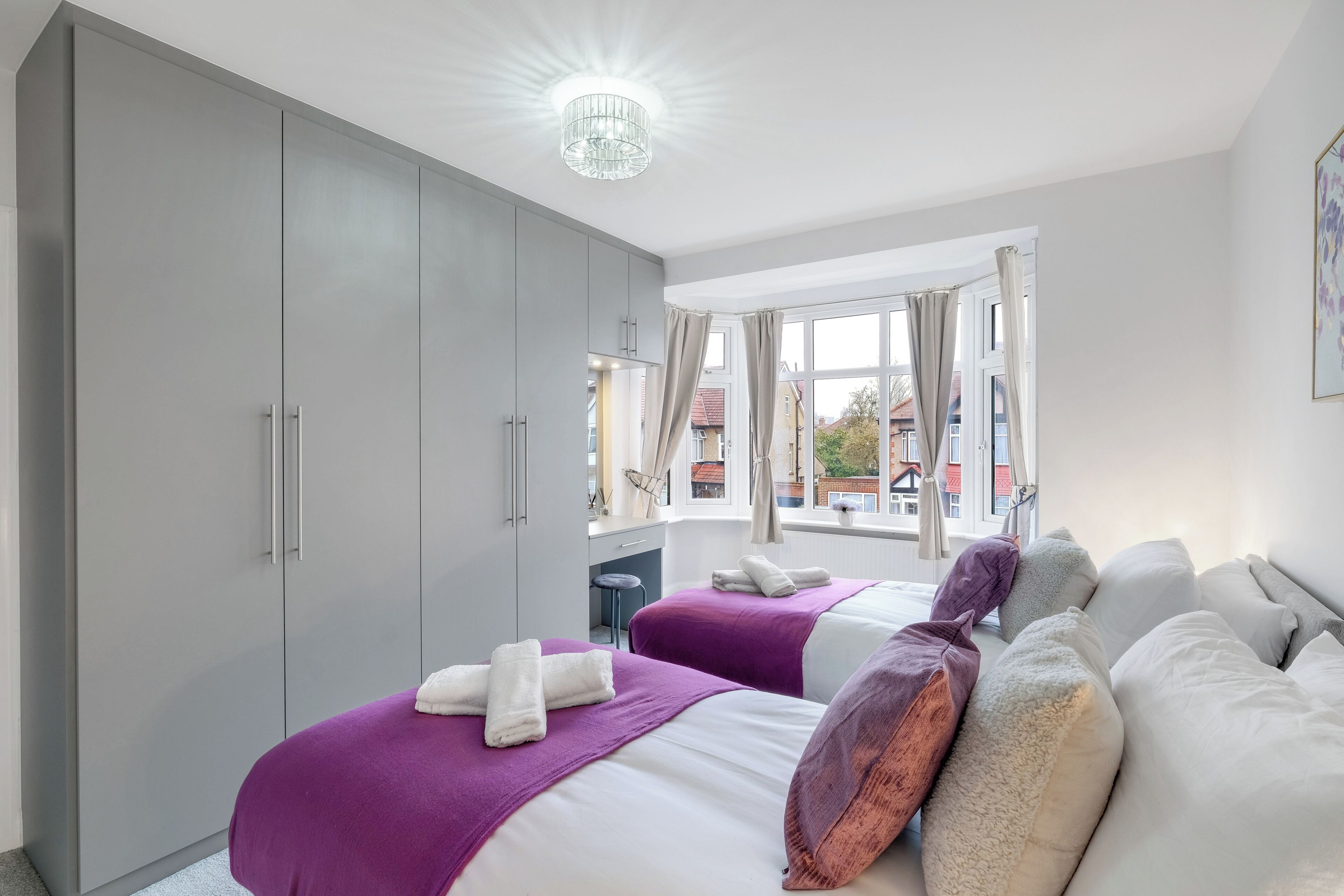 A hotel room with two beds, grey wardrobe, large window, and a chandelier.