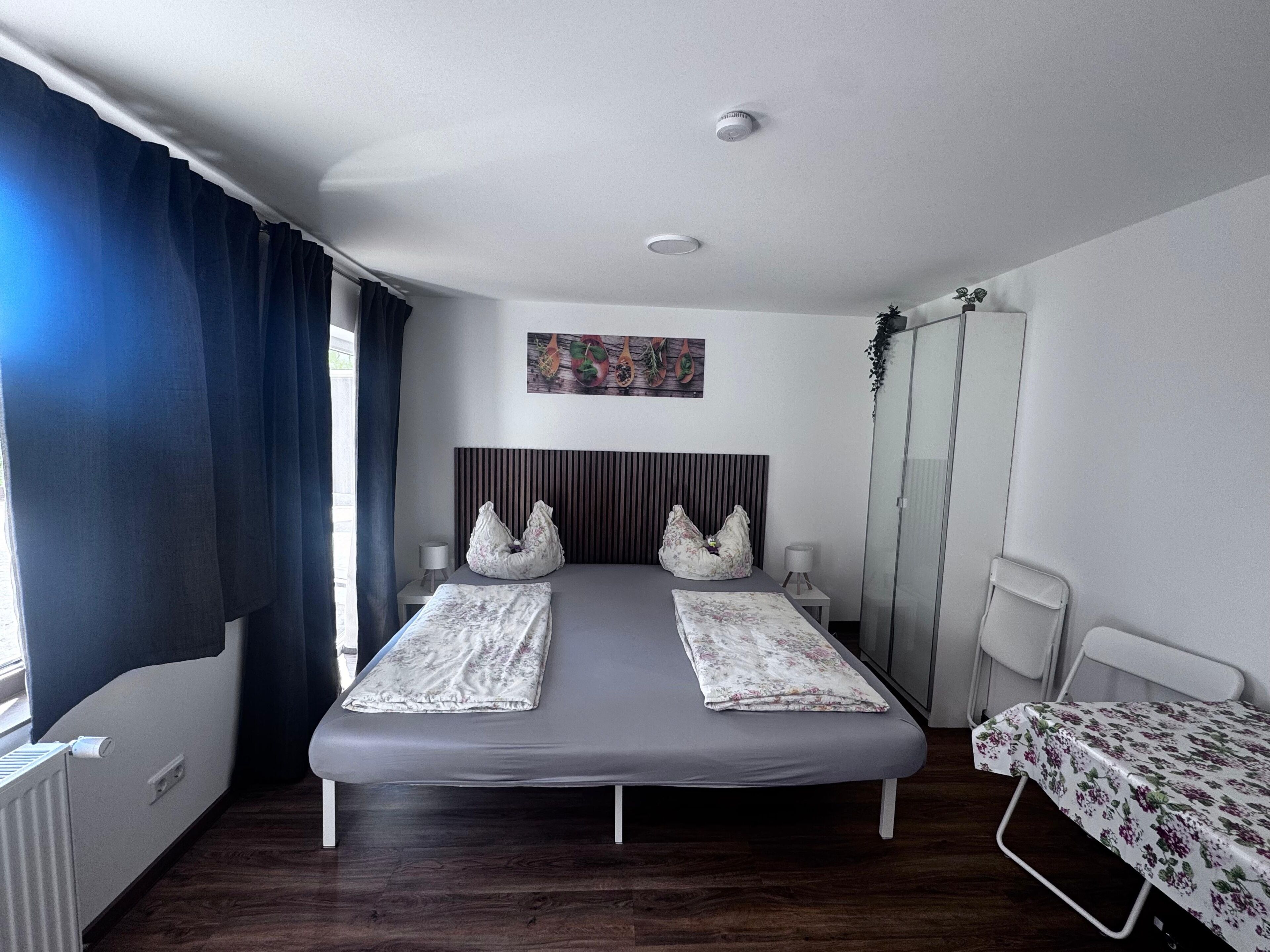 A double bed with a headboard, two bedside tables, a window with curtains, a radiator, and a wall-mounted artwork.