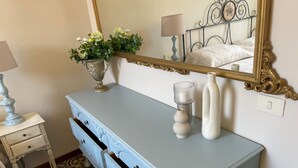 A room with a light blue dresser, a mirror, a vase with flowers, a lamp, and a patterned carpet. - Casa Mariuccia – 3 Bedrooms – In the Heart of Lucca (Lucca)