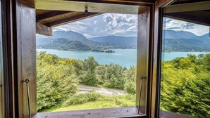Ventana de madera con vista a un lago, montañas y vegetación.