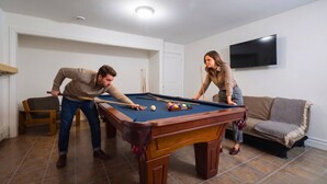 Une table de billard avec des billes, un canapé et un téléviseur dans une pièce aux murs blancs et au sol carrelé.