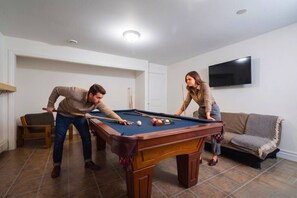 Une table de billard avec des billes, un canapé et un téléviseur dans une pièce aux murs blancs et au sol carrelé.
