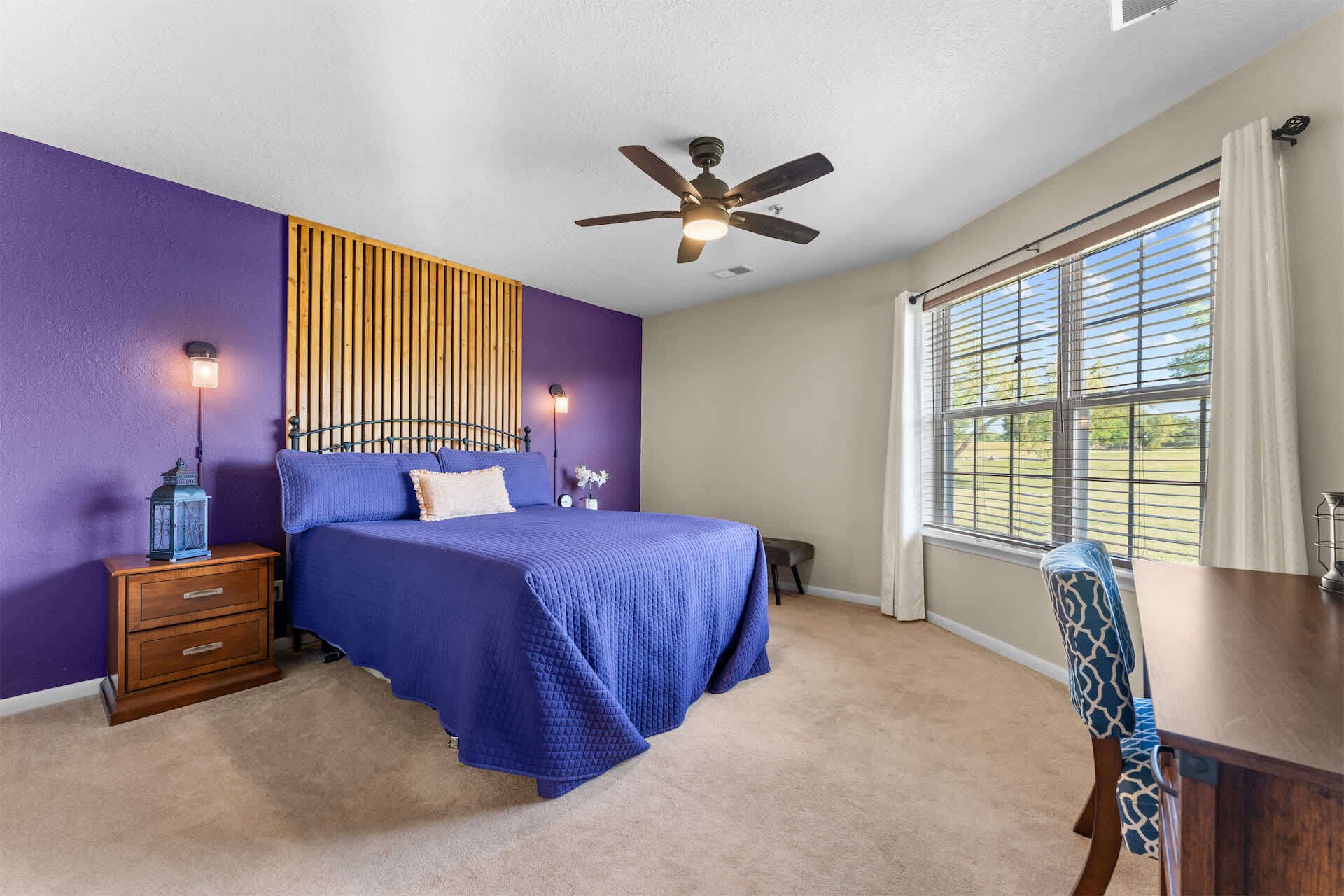 Une chambre à coucher comprenant un lit, une table de chevet, une lampe, un ventilateur de plafond, une fenêtre et une chaise.