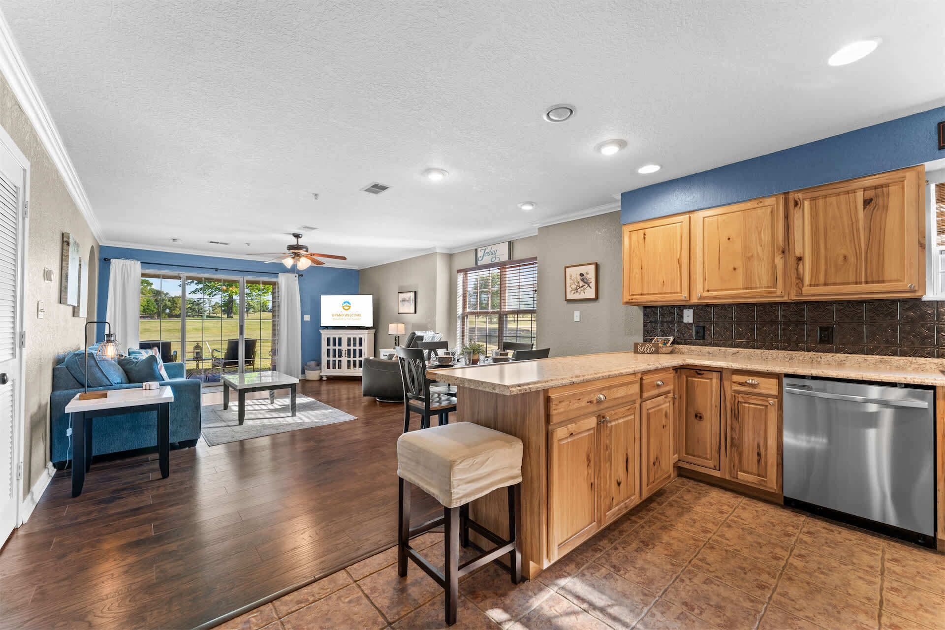 A spacious living area with a large TV, a dining table, and a kitchen with wooden cabinets and a stainless steel dishwasher.