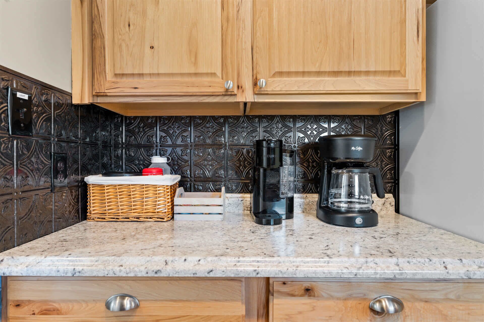 Una cocina con armarios de madera, encimera de mármol, cafetera, botella de agua y cesta de mimbre.