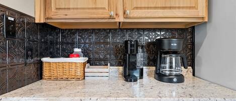 Una cocina con armarios de madera, encimera de mármol, cafetera, botella de agua y cesta de mimbre.
