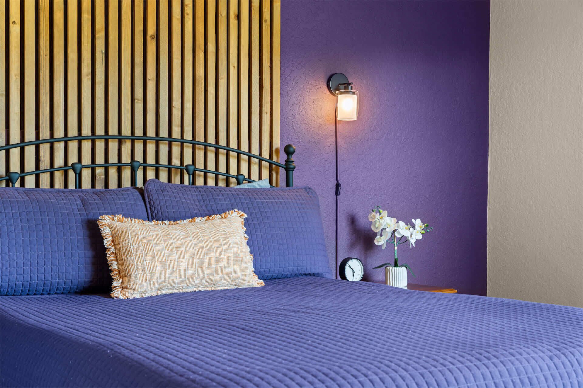 A bed with a headboard, a wall-mounted light, a clock, and a vase with flowers.