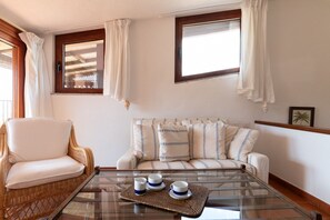 Ein Zimmer mit einem Korbstuhl, einer Couch mit gemusterten Kissen, einem Glastisch mit Untersetzer und zwei Fenstern mit Vorhängen.