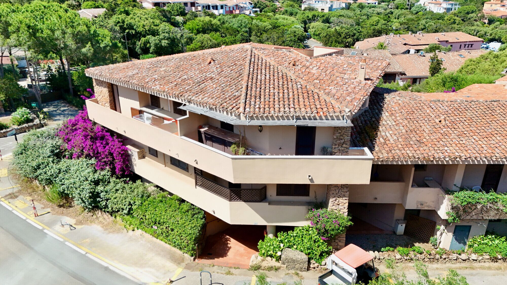 A multi-story building with a tiled roof, balconies, and a garden with purple flowers.