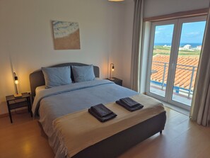 A bedroom with a bed, bedside tables, a nightstand, a window with a view of the sea, and a painting on the wall. - Villa Chaka | 4BR | Ocean View (Peniche)