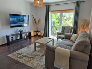 Ein Wohnzimmer mit Sofa, Couchtisch, Fernseher und Blick auf Grün durch das Fenster.