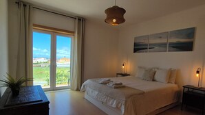 Ein Schlafzimmer mit einem großen Bett, zwei Nachttischen mit Lampen, einer Topfpflanze und Blick ins Freie.