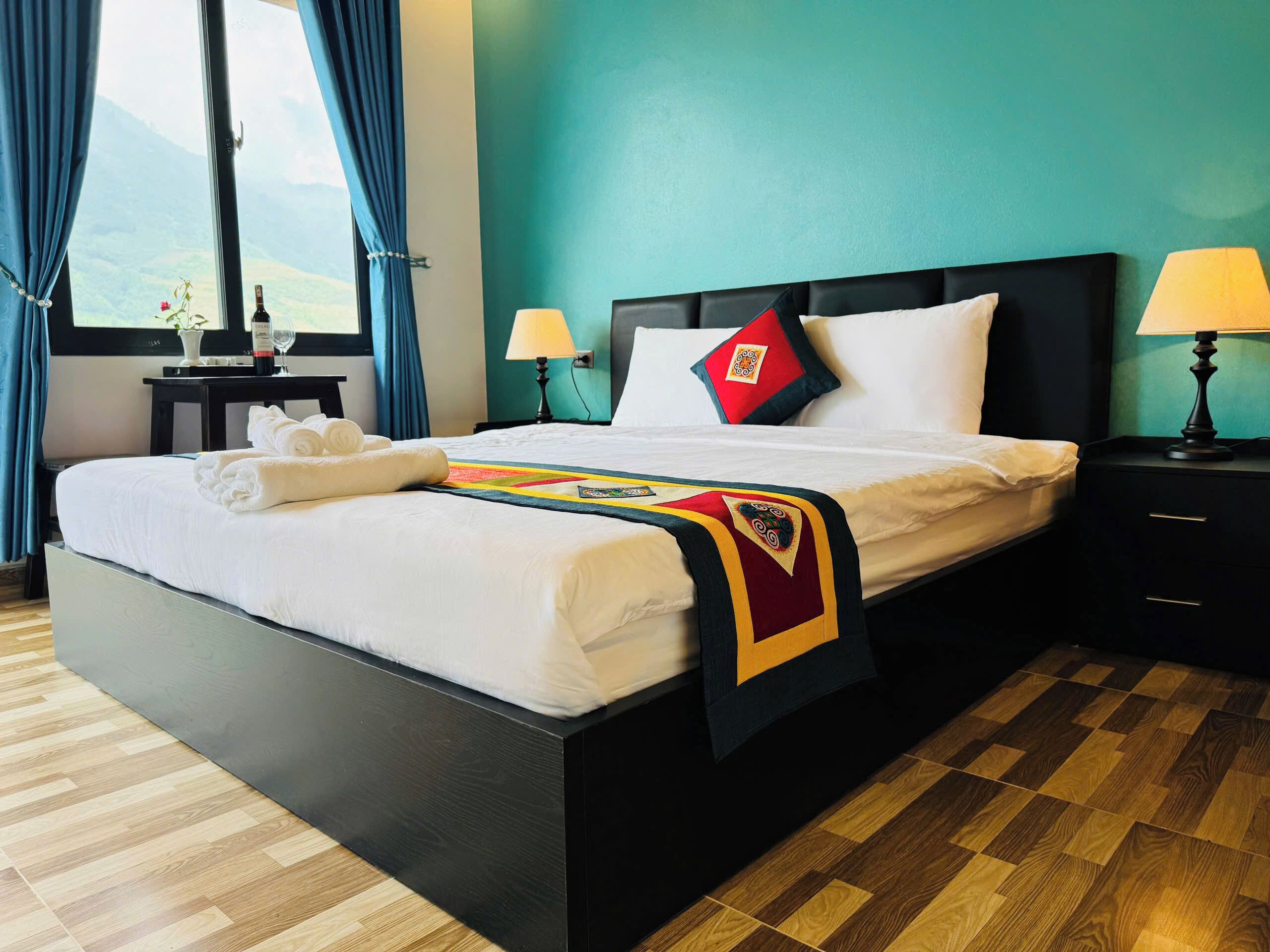 Una cama bien hecha con cobija y toallas, una mesita de noche con lámpara, y vista al exterior a través de una ventana.