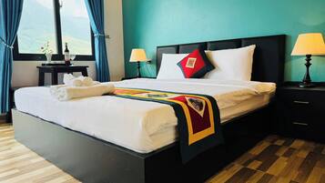 Una cama bien hecha con cobija y toallas, una mesita de noche con lámpara, y vista al exterior a través de una ventana.