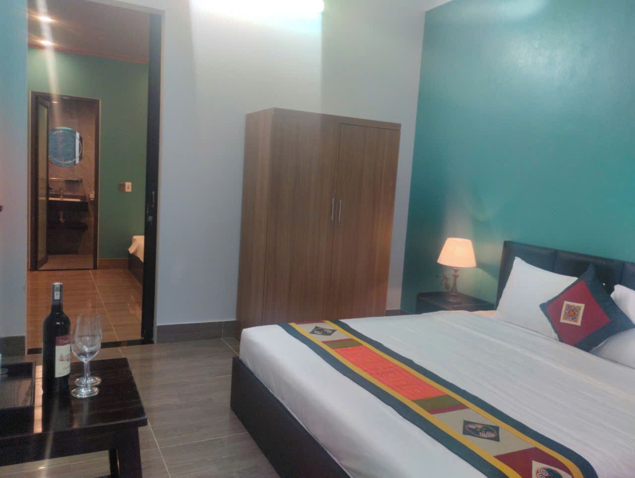 Habitación de hotel con cama, armario, mesita de noche con lámpara y una botella de vino sobre una mesa.