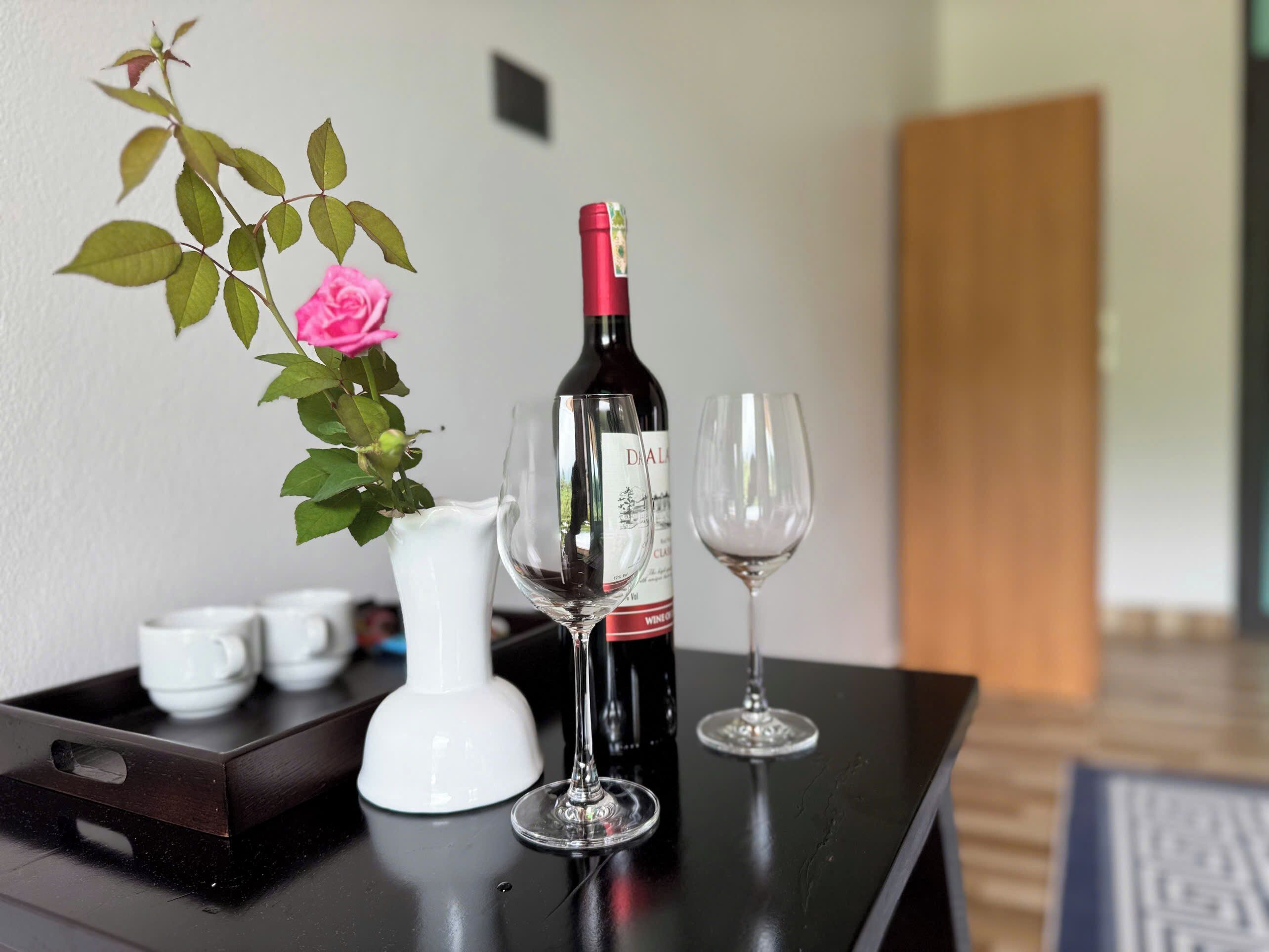 A bottle of wine, two wine glasses, a vase with a rose, and a tray with cups on a table.