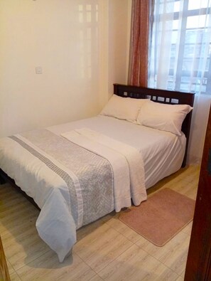 A single bed with a wooden headboard, a white bedspread, and a brown rug on a wooden floor. - Cozy 1 Bedroom apartment near JKIA and the SGR (Nairobi)