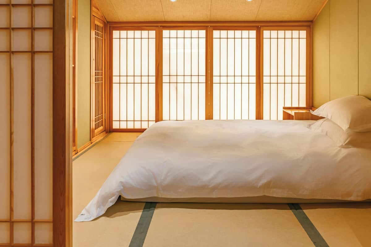 Ein traditioneller japanischer Raum mit Tatami-Boden, niedrigem Bett, Schiebetüren und Blick durch einen Shoji-Bildschirm.