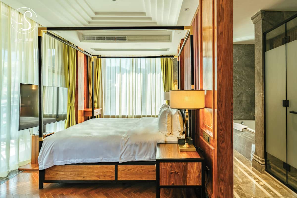 Ein Hotelzimmer mit Bett, Fernseher, Lampe und einem durch eine Glastür sichtbaren Badezimmer.