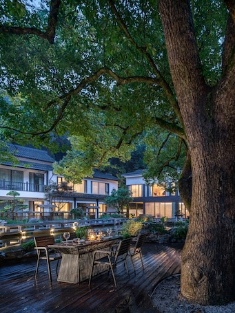 A serene outdoor dining area with a wooden deck, a table with chairs, and a large tree providing shade.. Manshe Creek Garden Boutique Resort