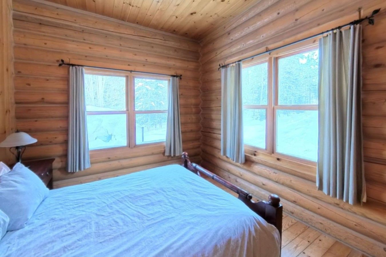A wooden cabin bedroom with a bed, two windows, and a lamp.