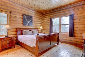 Une chambre à coucher dans une cabane en bois, avec un lit, deux tables de chevet, une lampe, un tableau et une fenêtre avec des rideaux.