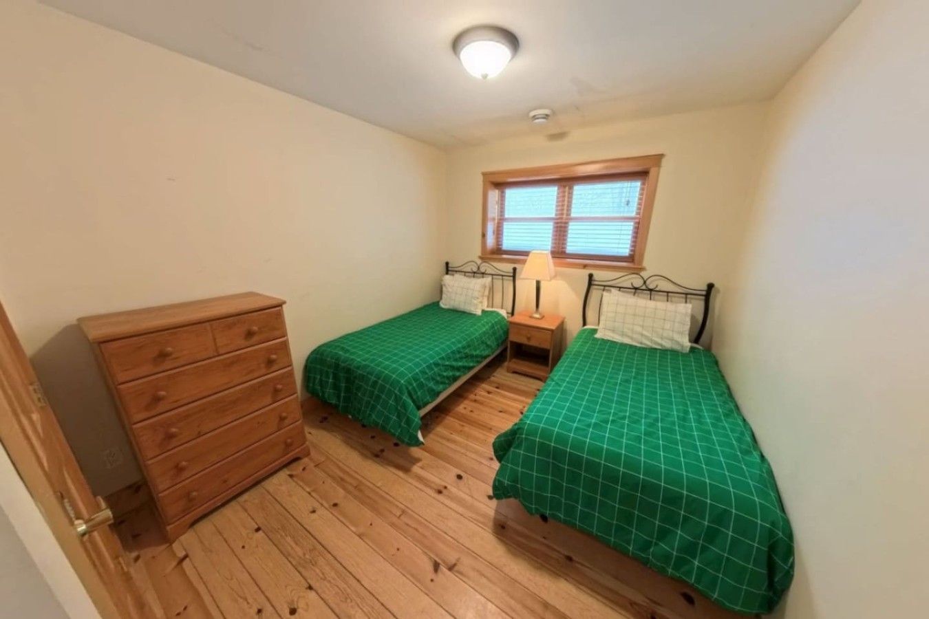 A small room with two single beds, a wooden dresser, a window with blinds, and a ceiling light.