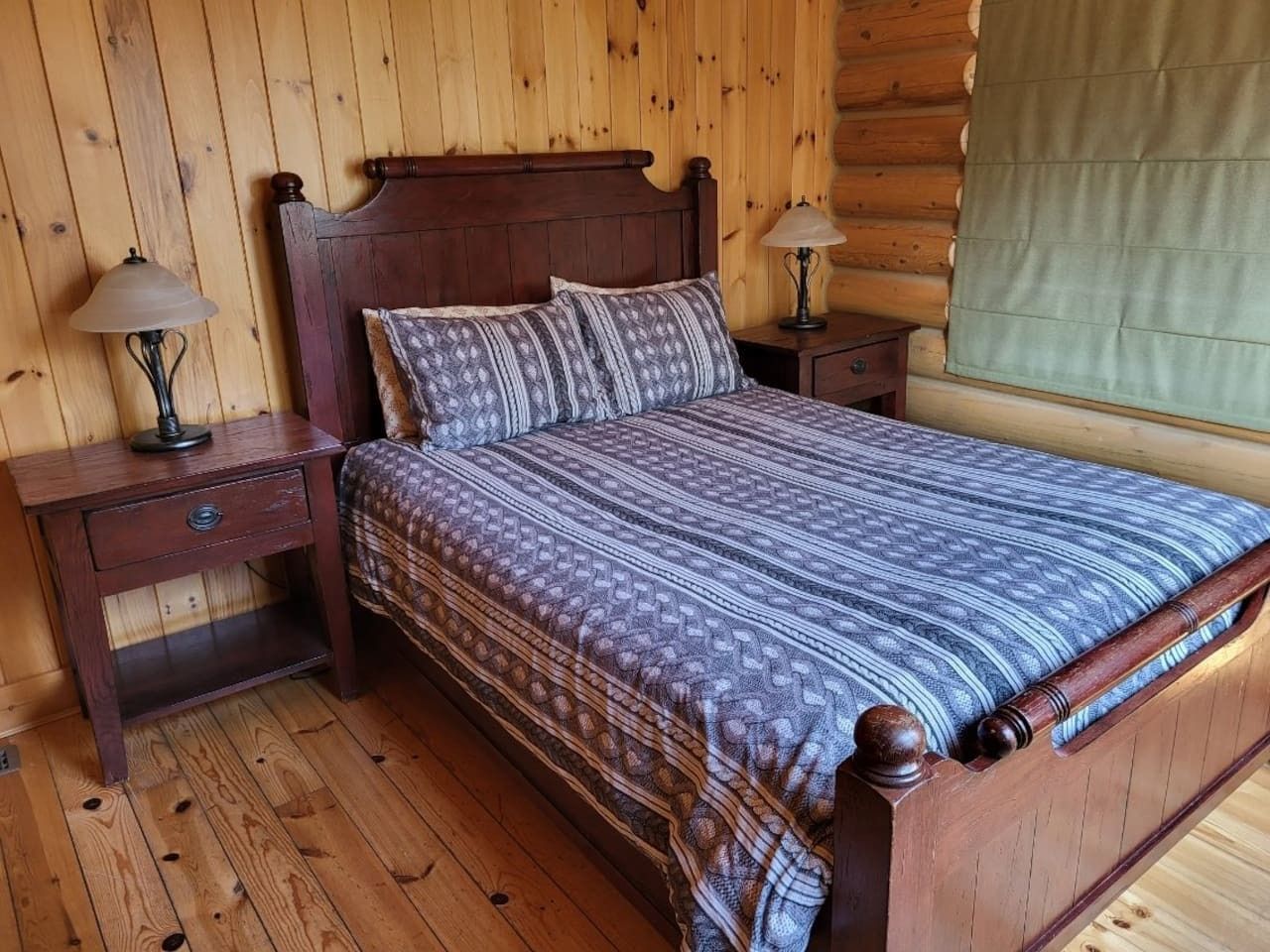 A wooden bed with a patterned bedspread in a room with wooden walls and a window with a green blind.